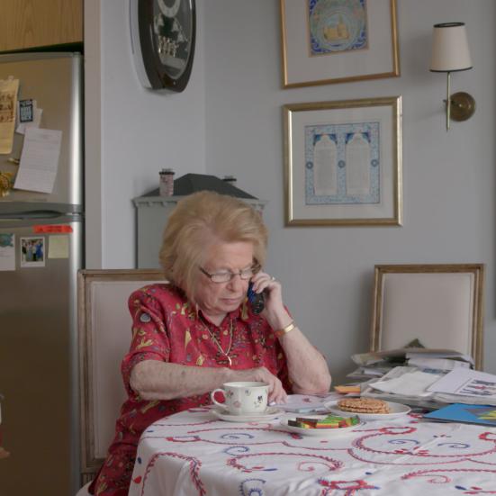 En ældre kvinde der sidder med en telefon mod sit øre ved et bord i et køkken